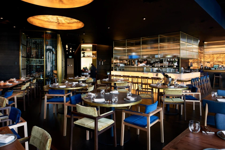 Upscale restaurant interior showcasing set tables with blue and yellow chairs, soft lighting, and bar area in the background.