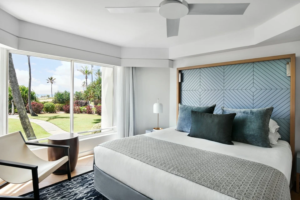 Bedroom interior featuring a king-size bed with blue accent pillows and headboard, chair, side table, and garden view from a large window.