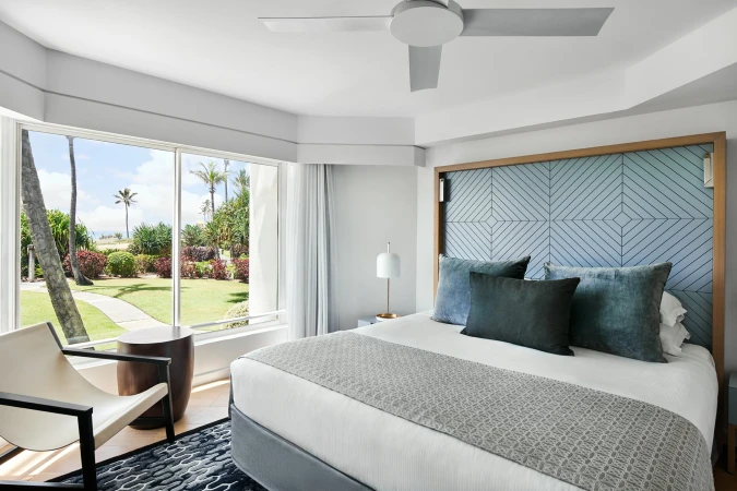 Bedroom interior featuring a king-size bed with blue accent pillows and headboard, chair, side table, and garden view from a large window.