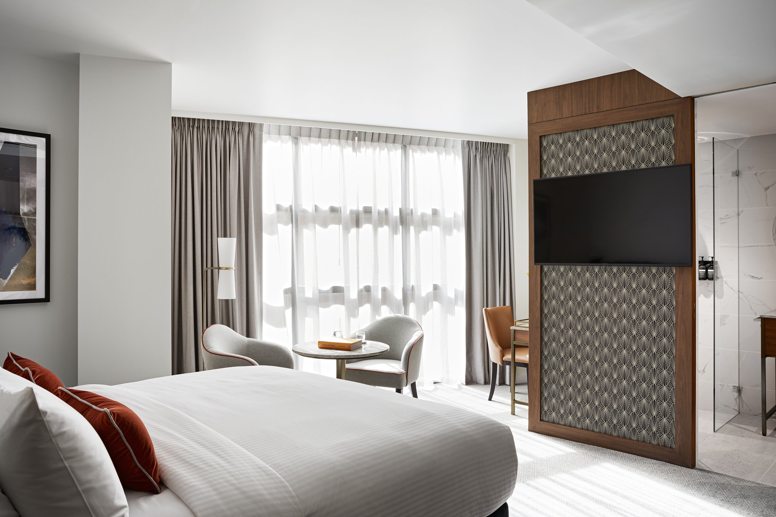 Hotel room interior with a king-size bed, seating area with a round table, and a mounted TV with patterned wall paneling.