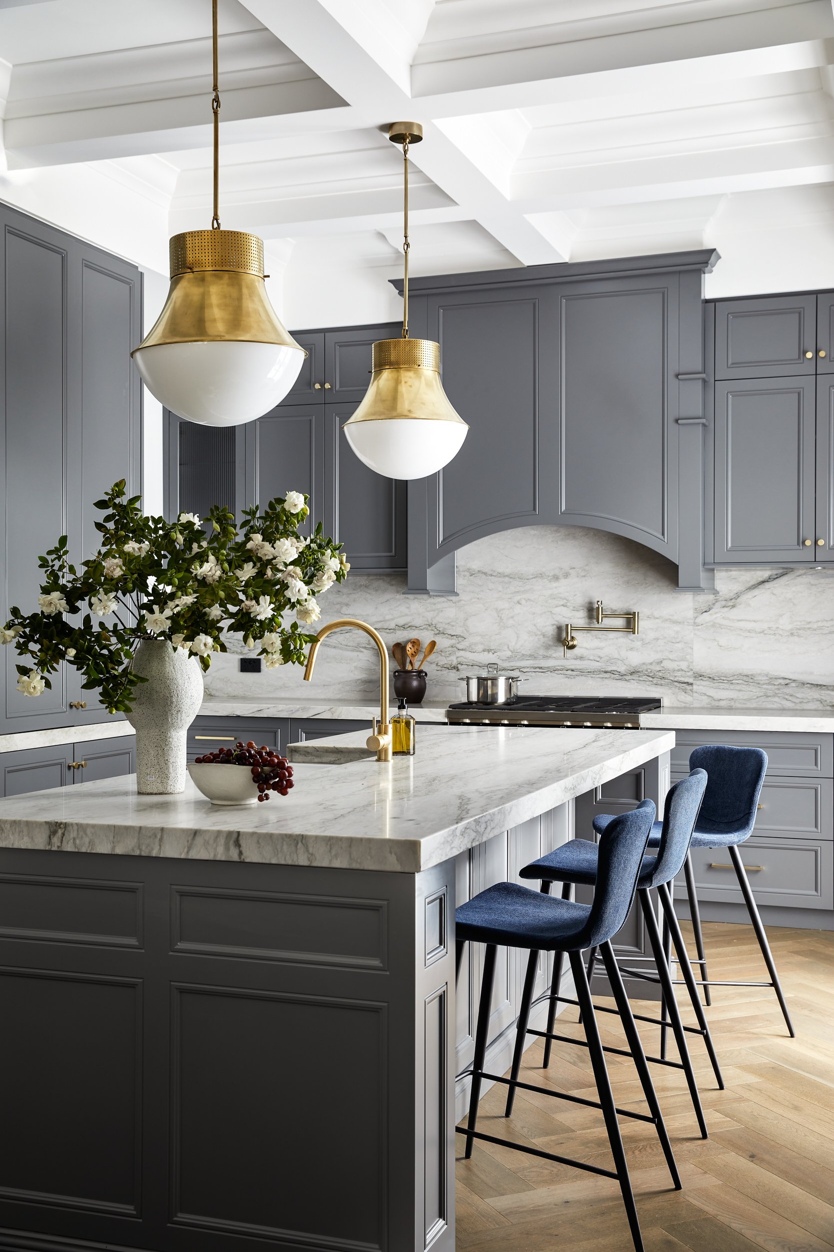 Bright kitchen with a marble island, navy blue bar stools, grey cabinets, gold hardware, and white and gold pendant lights.