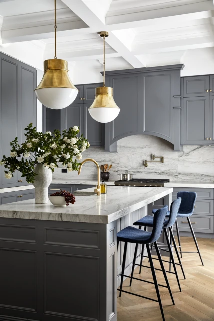 Bright kitchen with a marble island, navy blue bar stools, grey cabinets, gold hardware, and white and gold pendant lights.