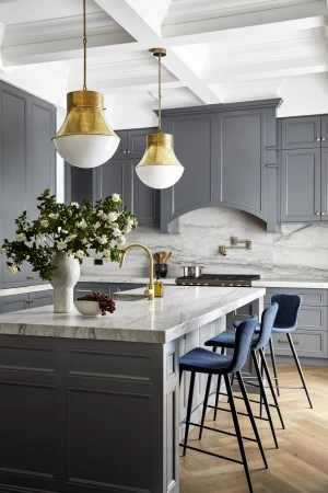 Bright kitchen with a marble island, navy blue bar stools, grey cabinets, gold hardware, and white and gold pendant lights.