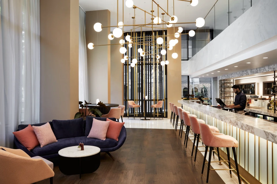 Interior of a stylish hotel lobby featuring a dark blue sofa with pink pillows, a marble bar with pink stools, and decorative lighting.