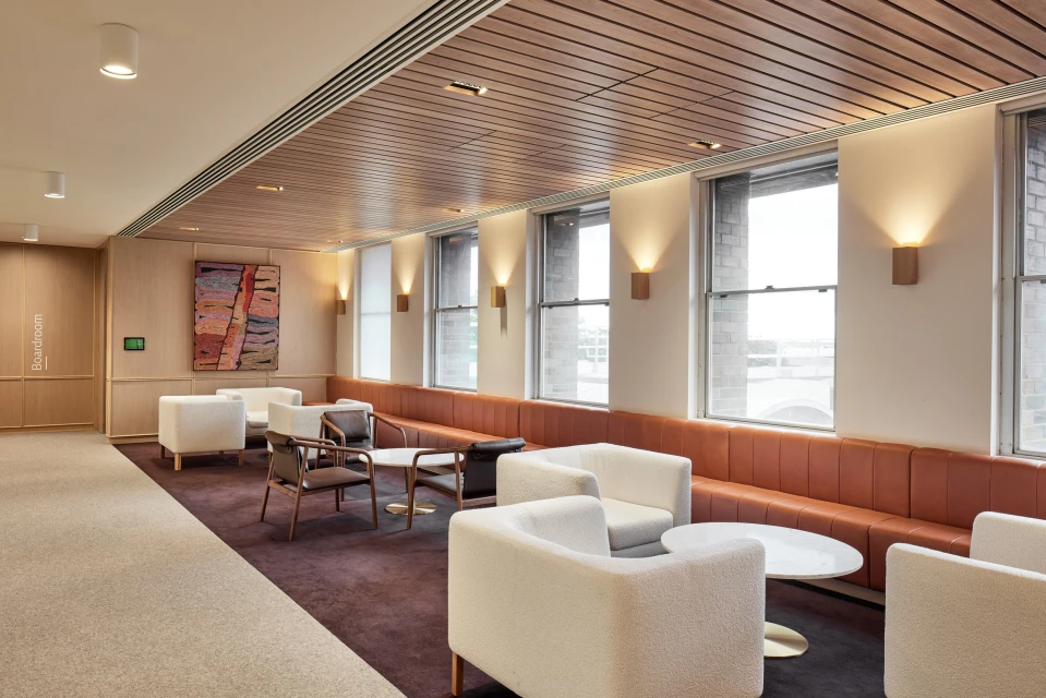 Modern office lounge with neutral-toned seating, wood ceiling, and large windows allowing natural light, creating an inviting space.