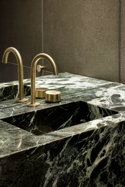 Close-up of a green marble countertop featuring golden brass fixtures, including two curved faucets and cylindrical handles.