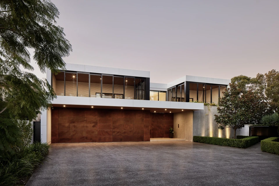 Exterior of a modern two-story house featuring a flat roof, floor-to-ceiling windows, and a dark wood garage door, set in a landscaped yard.