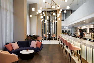Interior of a modern hotel bar with a marble counter, pink velvet stools, and a large multi-bulb golden chandelier.