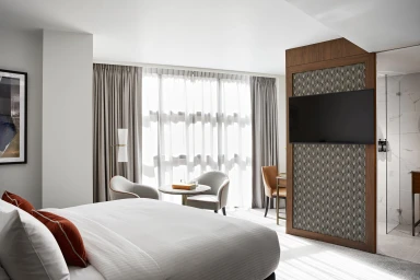Modern hotel room interior featuring a made bed with white linens and orange pillows, armchairs by the window, and a wall-mounted TV.
