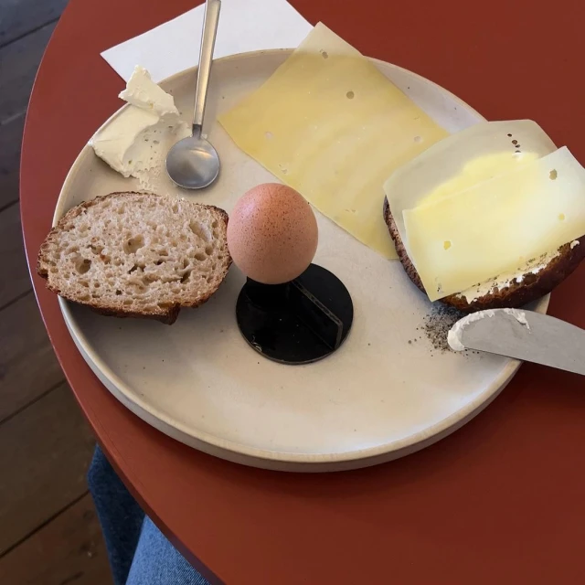Overhead shot of a plate with sliced bread, cheese, butter, and a boiled egg balanced on a black stand on a red table.