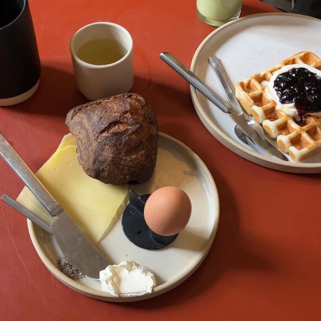 Overhead shot of a breakfast plate with bread, cheese, an egg, cream, and waffle with jam on a red surface. 