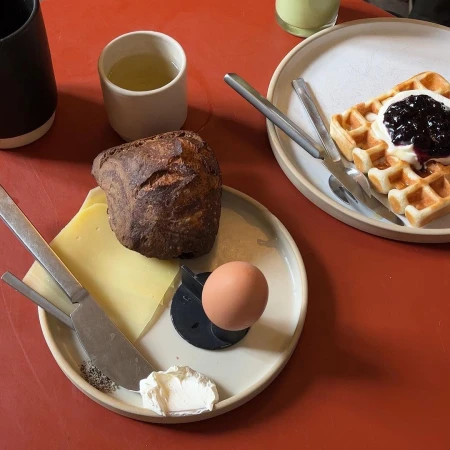 Overhead shot of a breakfast plate with bread, cheese, an egg, cream, and waffle with jam on a red surface. 