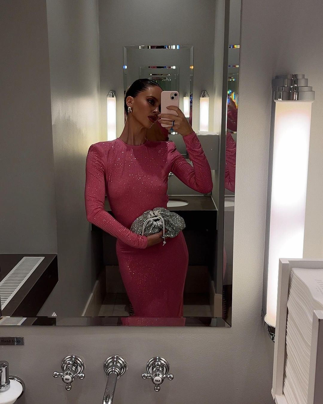 Elegant adult woman takes a selfie in a bathroom, wearing a long-sleeve pink dress and holding a silver clutch.
