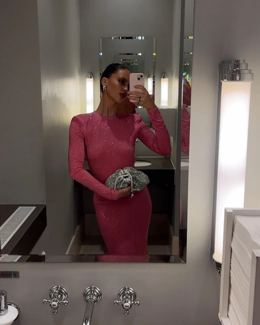 Elegant adult woman takes a selfie in a bathroom, wearing a long-sleeve pink dress and holding a silver clutch.