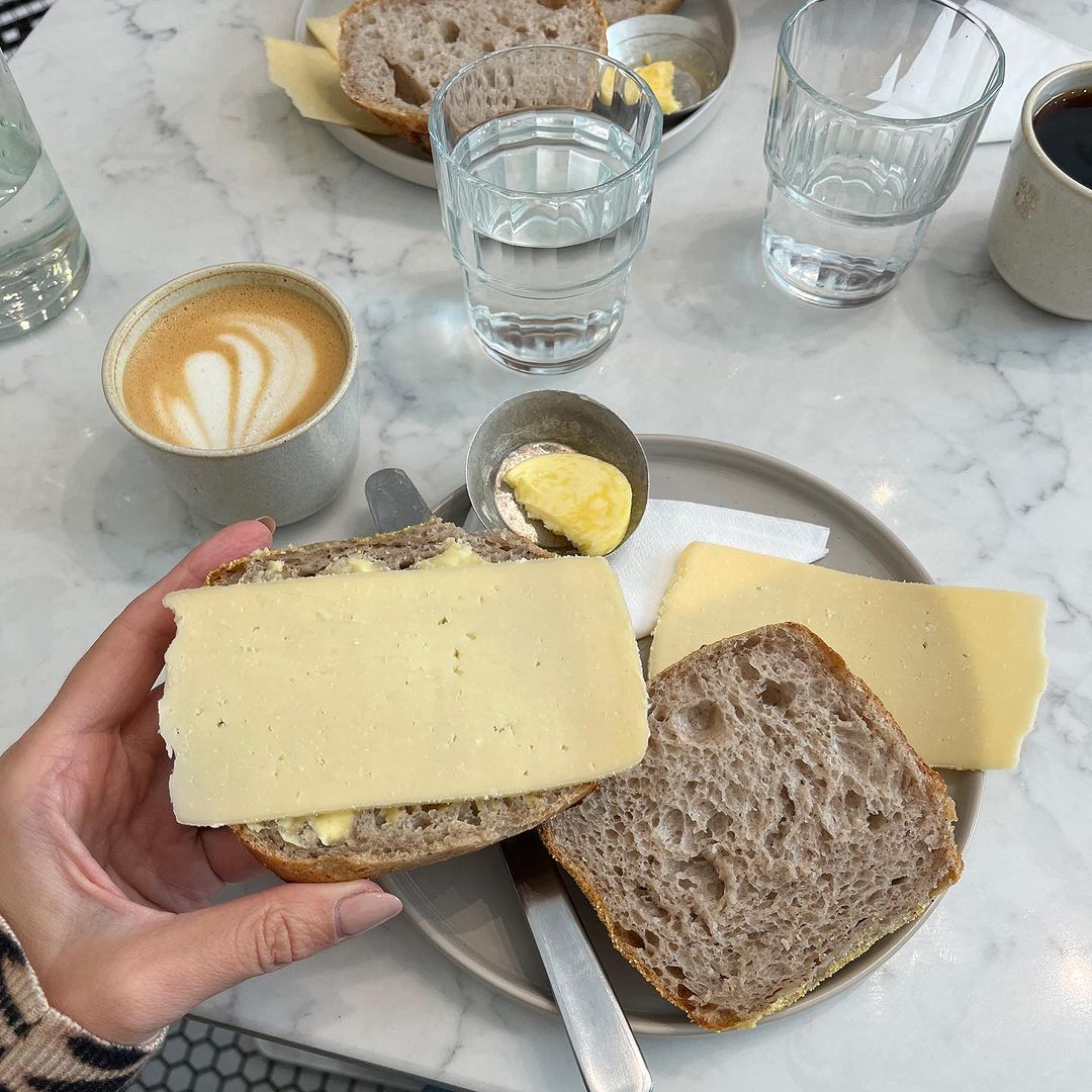 Overhead shot of a person holding a slice of bread topped with cheese, alongside coffee, water, and butter on a marble table.