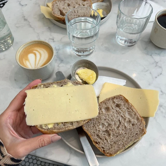 Overhead shot of a person holding a slice of bread topped with cheese, alongside coffee, water, and butter on a marble table.