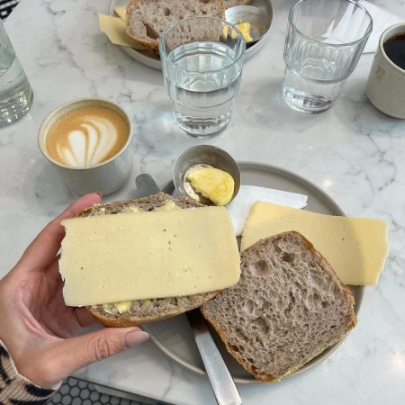 Overhead shot of a person holding a slice of bread topped with cheese, alongside coffee, water, and butter on a marble table.