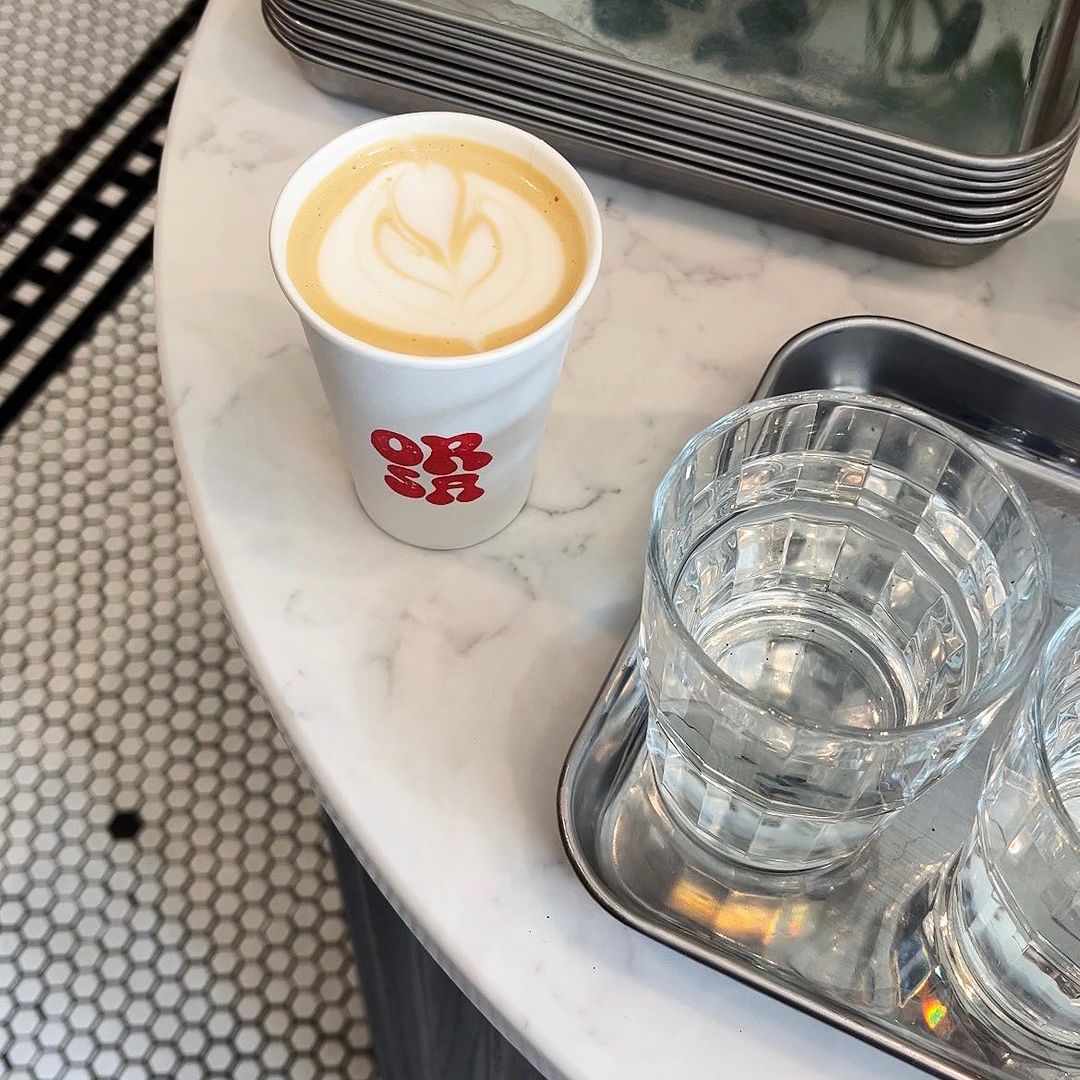 Overhead shot of a latte with latte art in a paper cup on a marble table next to a glass of water and a metal tray.