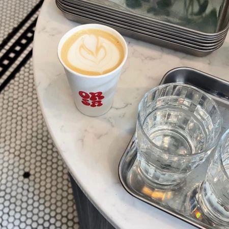 Overhead shot of a latte with latte art in a paper cup on a marble table next to a glass of water and a metal tray.