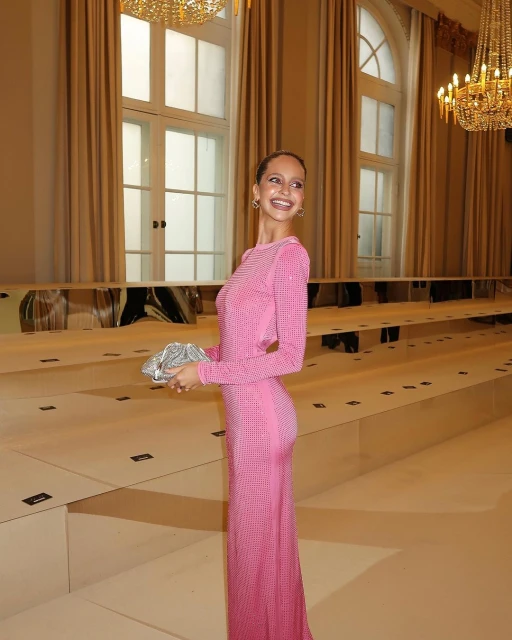 Elegant woman in a pink gown holding a silver clutch, standing inside a room with beige walls, large windows and a decorative chandelier.