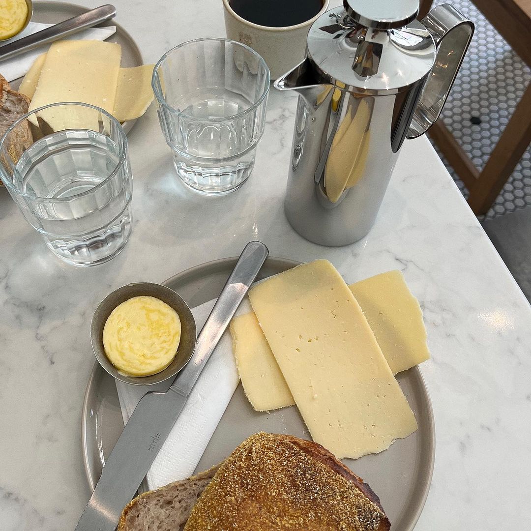 Overhead shot of a marble table with breakfast items: cheese slices, bread, butter, coffee in a mug, and a french press coffee maker.