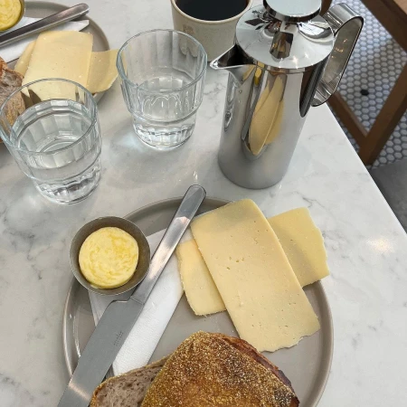 Overhead shot of a marble table with breakfast items: cheese slices, bread, butter, coffee in a mug, and a french press coffee maker.