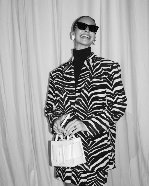 Smiling woman in a zebra-print suit, dark sunglasses, and white earrings holds a small white purse against a draped background.