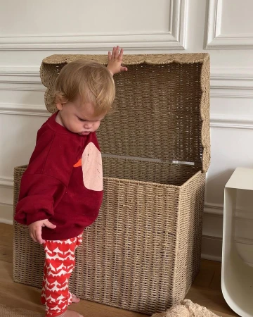 Toddler girl in patterned leggings reaches for open wicker toy chest in indoor setting with white wall and natural light.