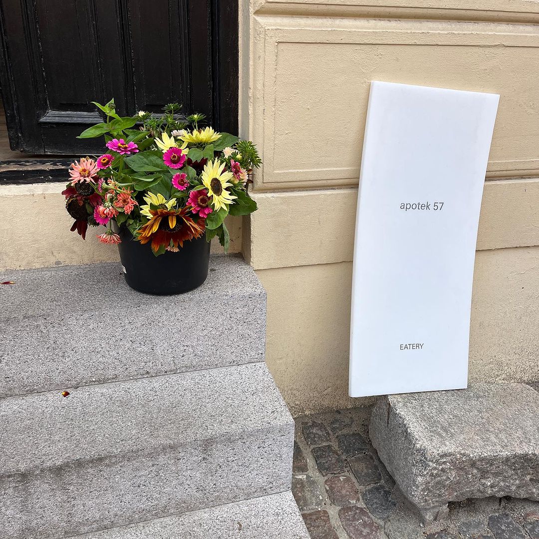 Outdoor shot of a colorful flower arrangement in a black pot on steps, next to a white sign for Apotek 57 Eatery.