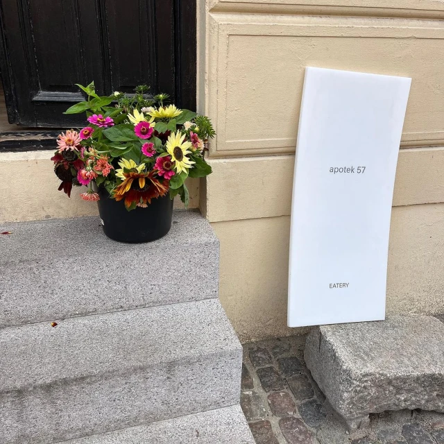 Outdoor shot of a colorful flower arrangement in a black pot on steps, next to a white sign for Apotek 57 Eatery.