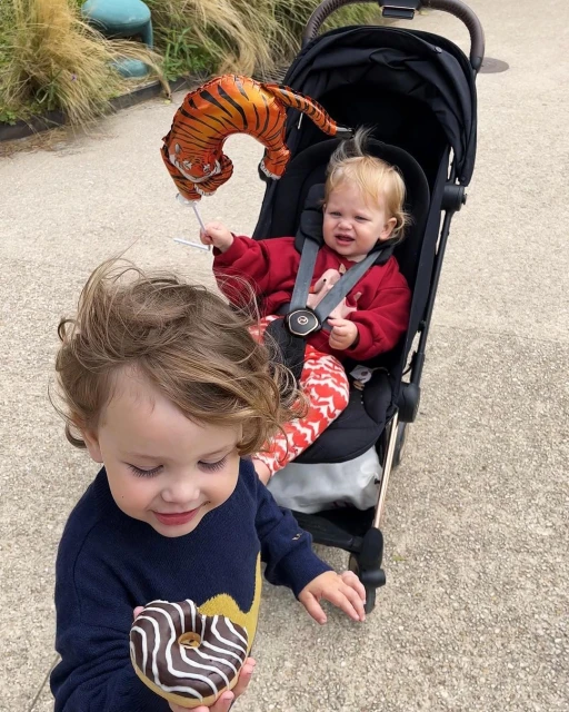 Two young children outdoors: one sitting in a stroller with a tiger balloon and another toddler holding a chocolate frosted donut.