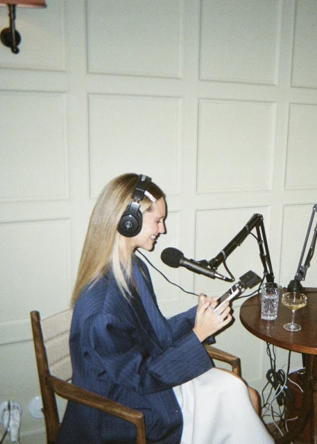 Woman recording podcast in a studio wearing headphones, holding phone, table with microphone, glass of water and wine in the frame.