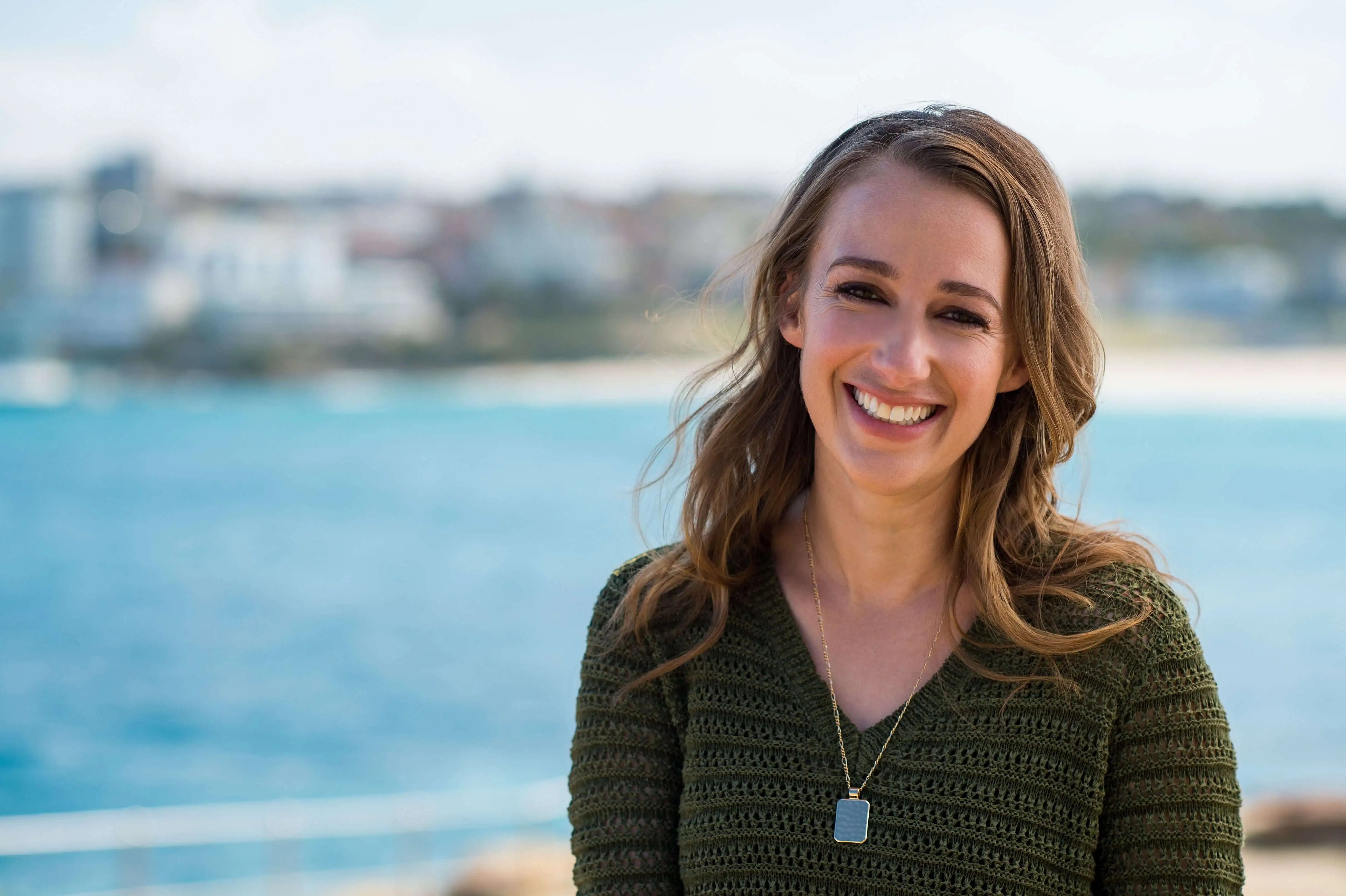 Smiling woman with long brown hair poses outdoors near the ocean. She wears a green sweater and gold necklace in medium shot.