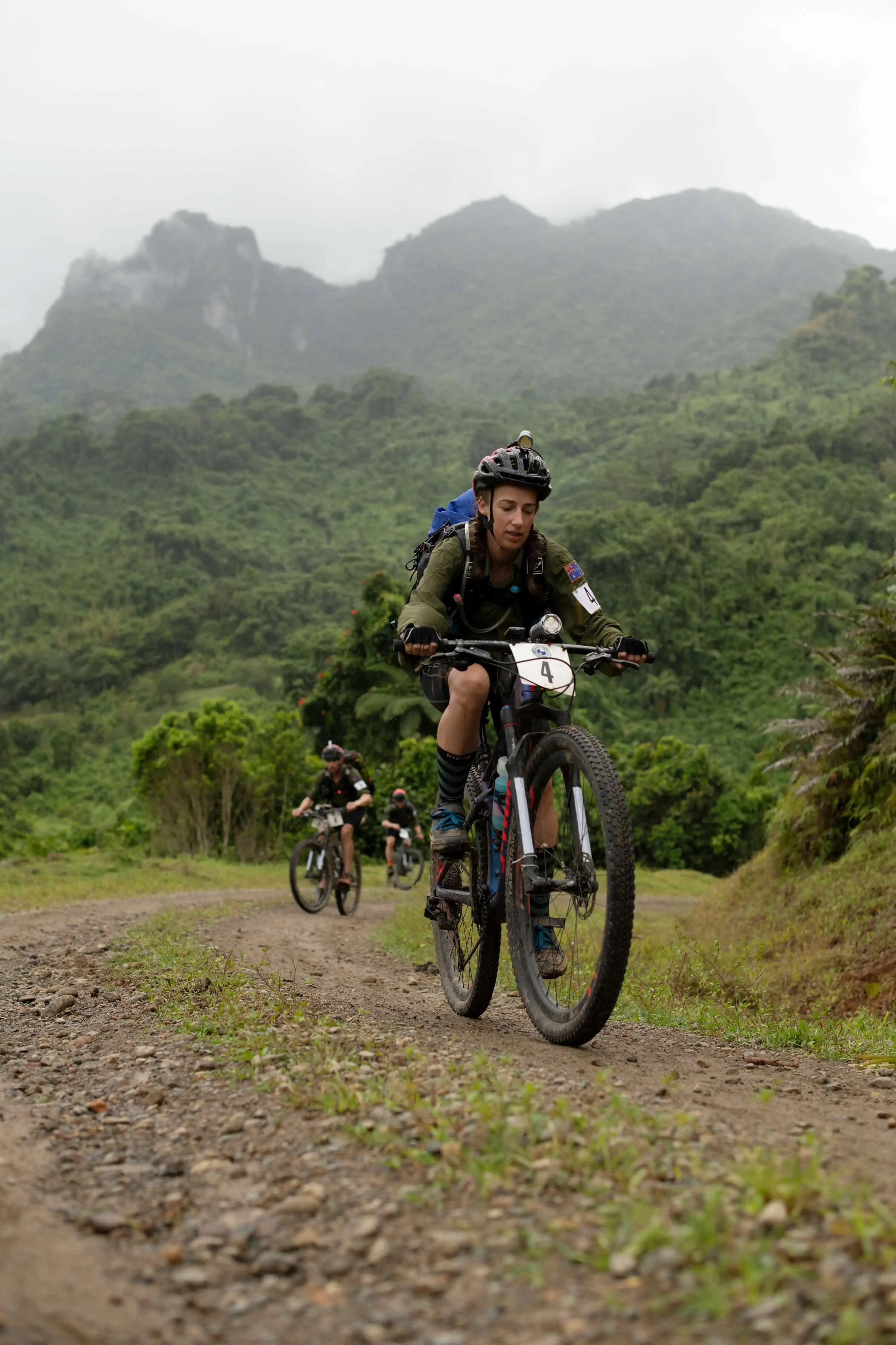 A group of mountain bikers rides on a dirt trail through a green mountainous landscape under an overcast sky, showcasing outdoor adventure.