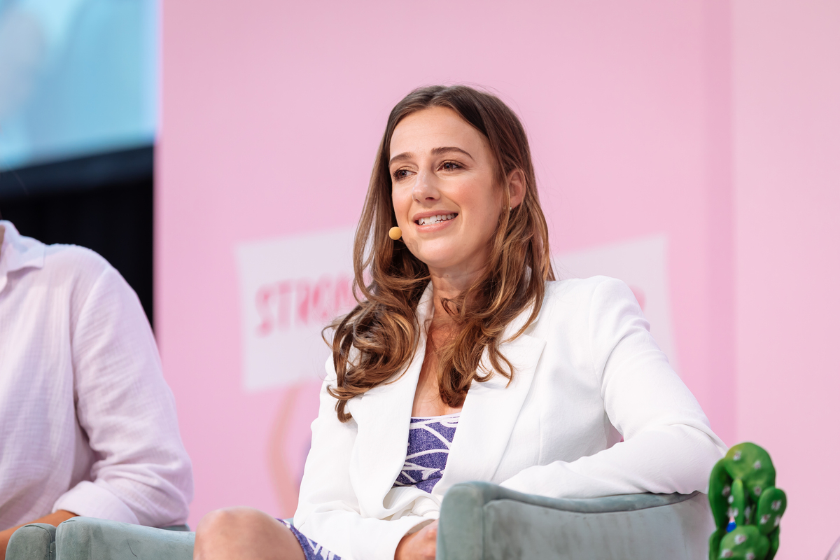 Woman in white blazer sits on stage with pink backdrop. She has brown hair and is smiling at the audience at an event.