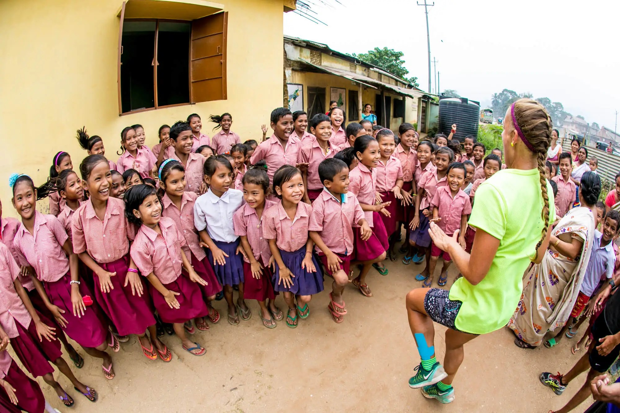 Cheerful elementary school students in matching uniforms gather around a female instructor in an outdoor setting with surrounding buildings.