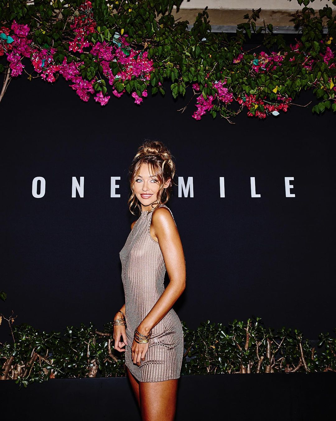 An adult woman poses in a shimmering dress with flowers in the background and 'ONE MILE' text. Her hair is styled in an updo.