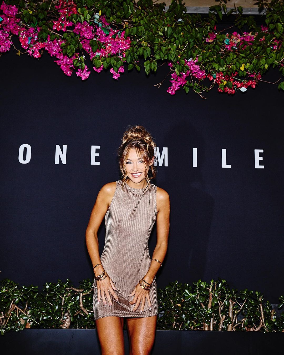 An adult woman stands in a gold dress in front of a black wall with a white sign, floral garlands, and green bushes beneath the sign.