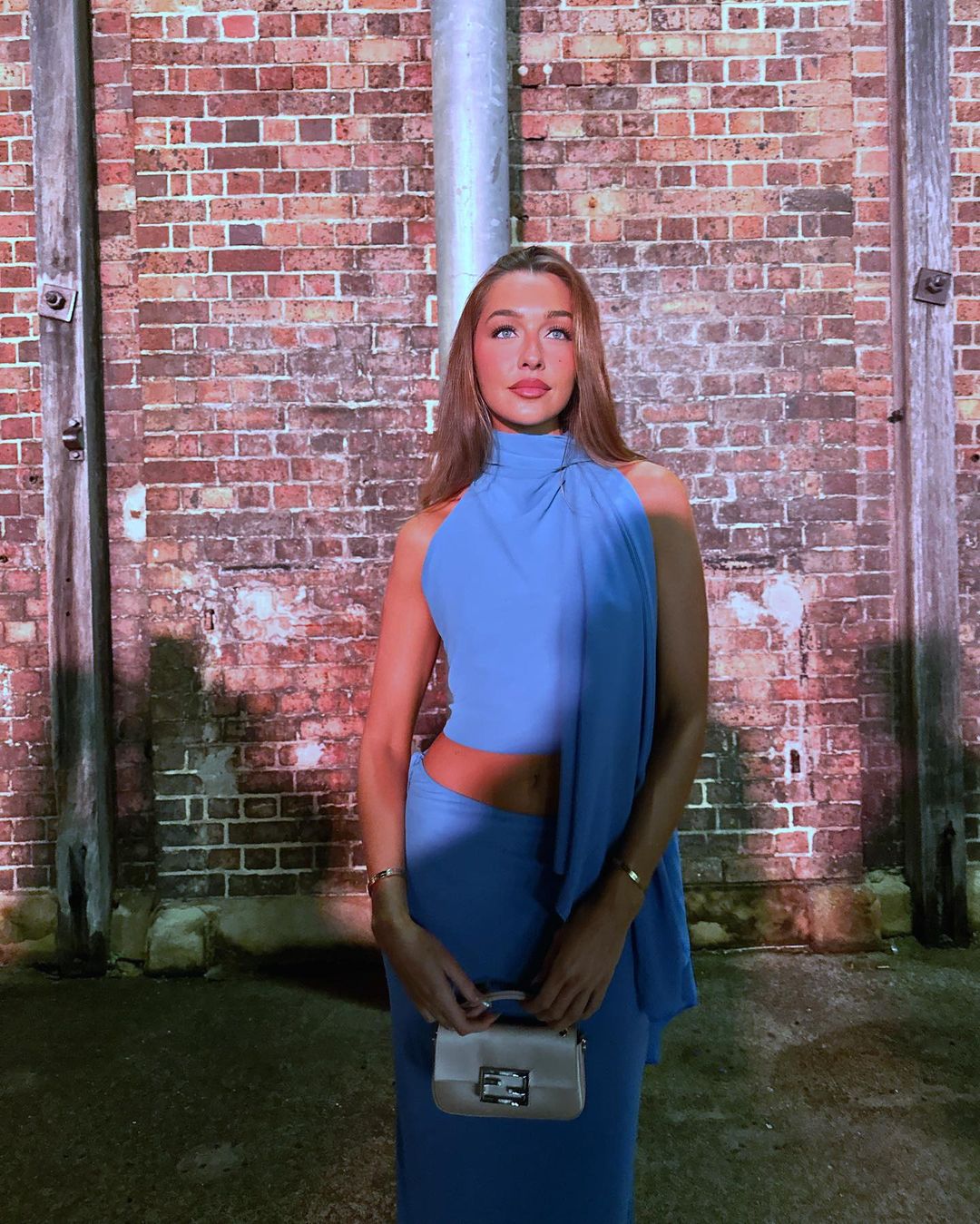 Woman poses against brick wall in blue halter dress with shoulder drape, accessorized with gold bracelet and small handheld purse.