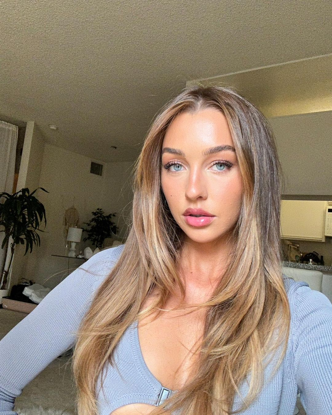 Portrait of a blonde woman with blue eyes, wearing a grey top, posing in a neutral indoor environment with a glimpse of plants in the background.