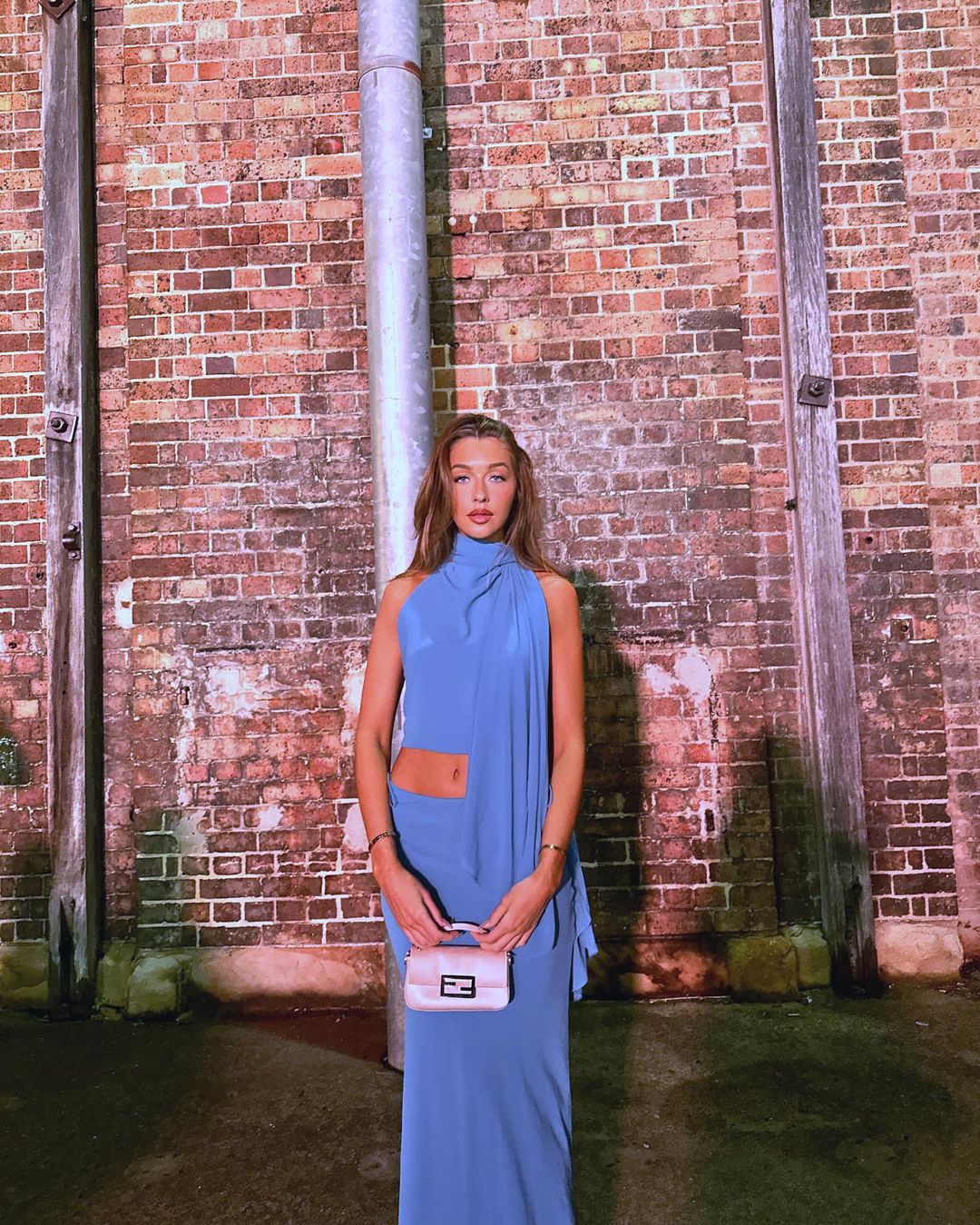 Adult woman in a blue halter dress and cream handbag stands in front of a red brick wall with a metal pipe fixture.