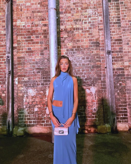 Adult woman in a blue halter dress and cream handbag stands in front of a red brick wall with a metal pipe fixture.