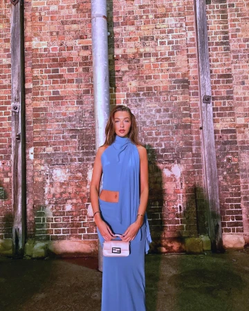 Adult woman in a blue halter dress and cream handbag stands in front of a red brick wall with a metal pipe fixture.