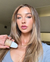 Portrait of a woman with long blonde hair holding a small foundation bottle, wearing a blue top against an interior background