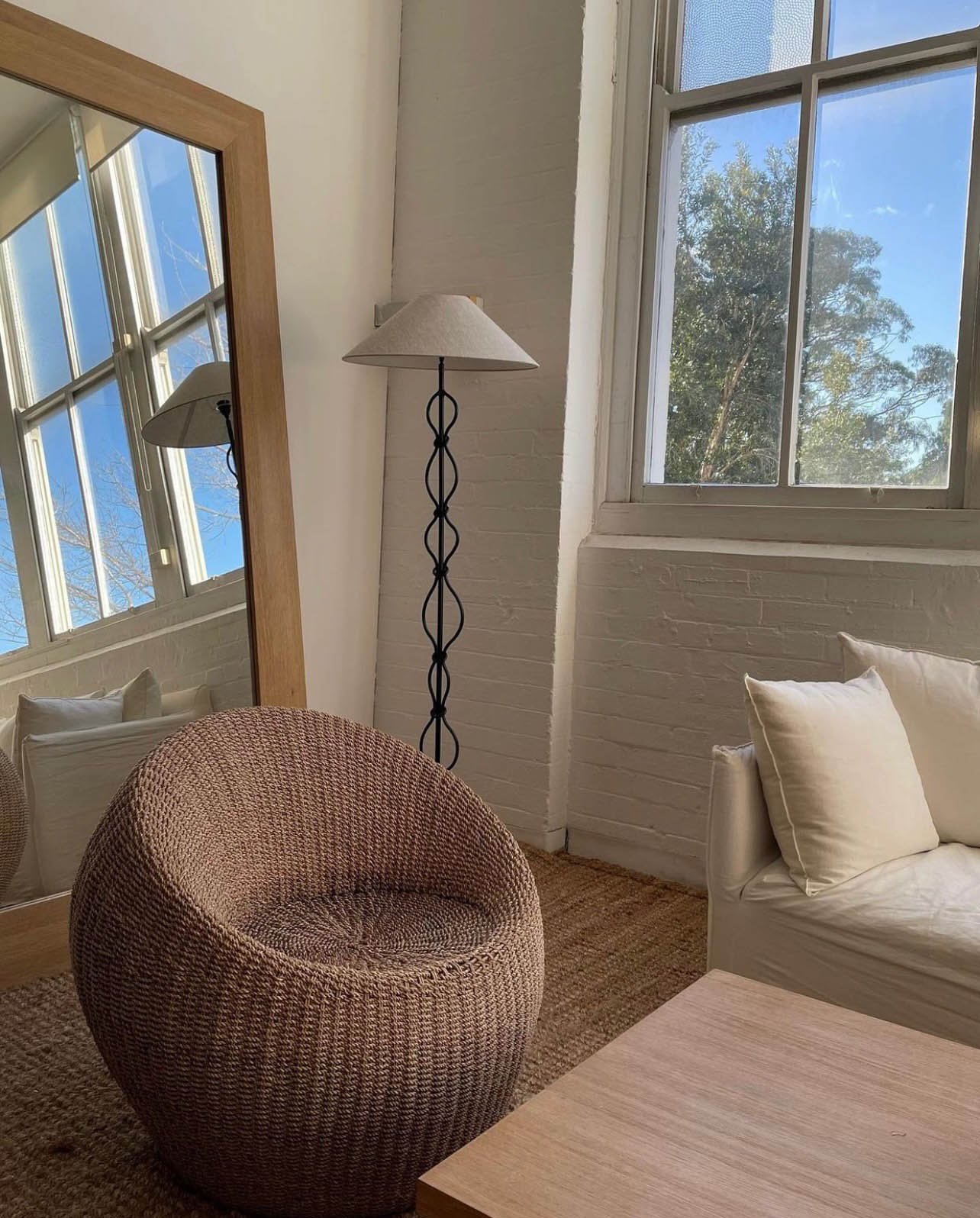 Interior scene with wicker chair, floor lamp, light wood furniture, and a large mirror reflecting the room, bathed in natural light.