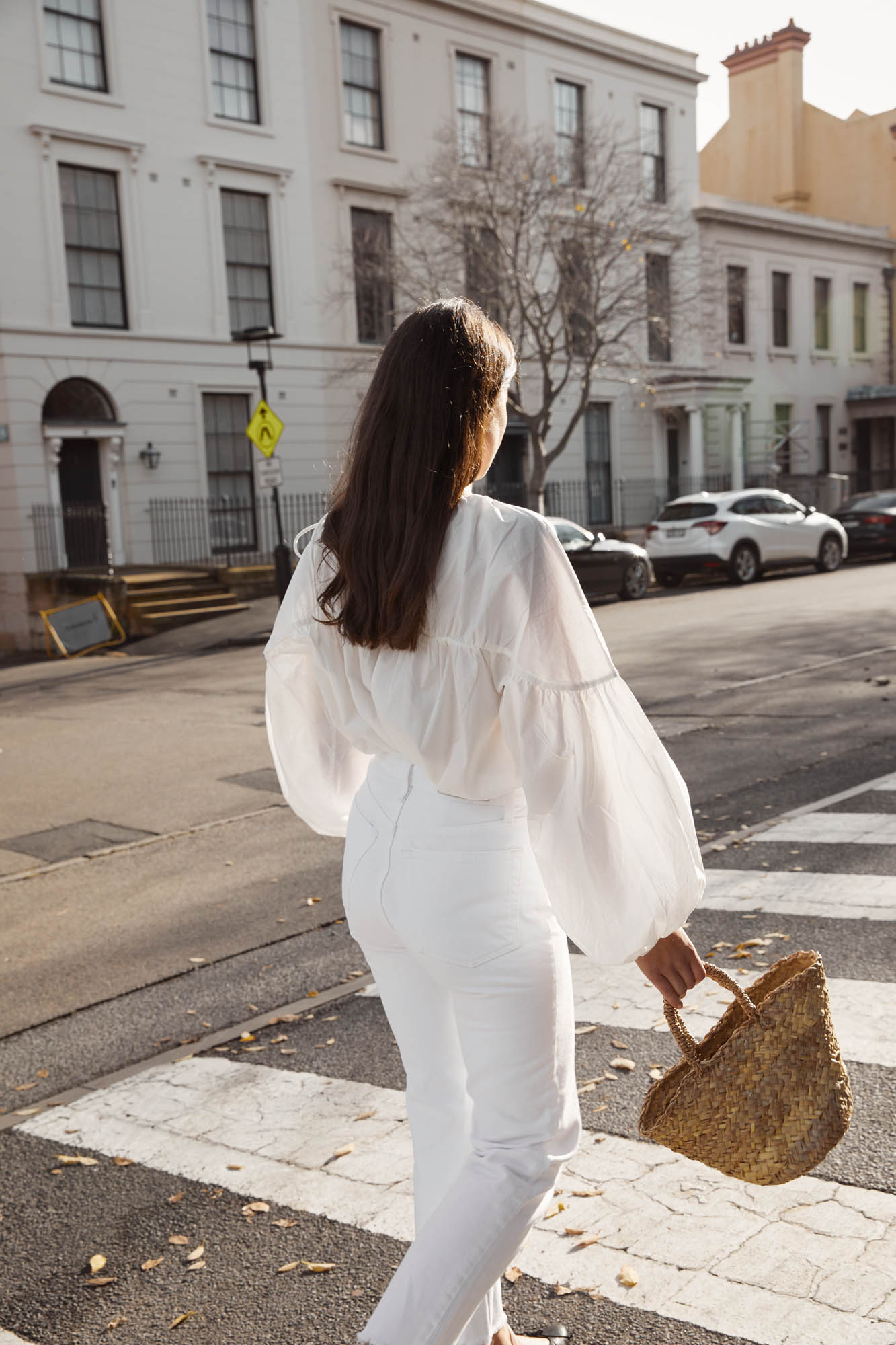 Woman in white crossing a street with residential buildings in the background, carrying a woven handbag on a sunny day.