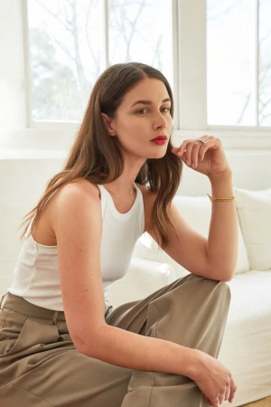Woman wearing a white tank top and grey trousers seated indoors against a light background with a white sofa
