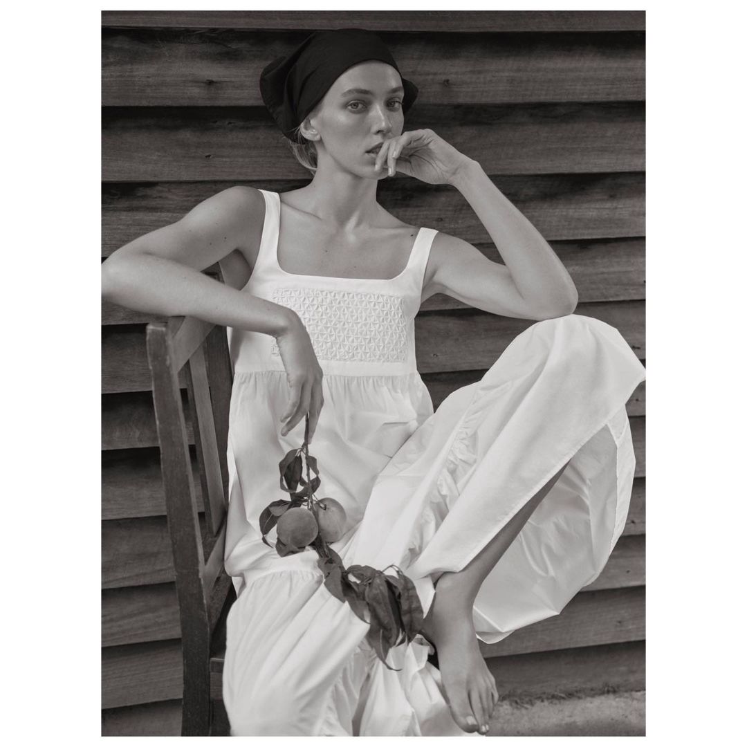 A woman in a white dress and headscarf sits on a wooden chair against a wooden wall, holding fruit and leaves in a monochromatic portrait.