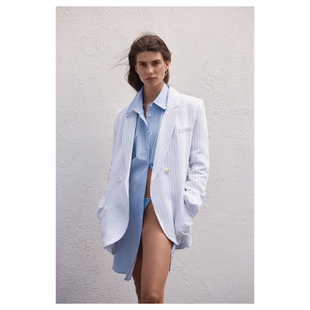 An adult female fashion model poses against a light-colored wall, wearing a blue button-down shirt and an oversized pinstripe blazer.