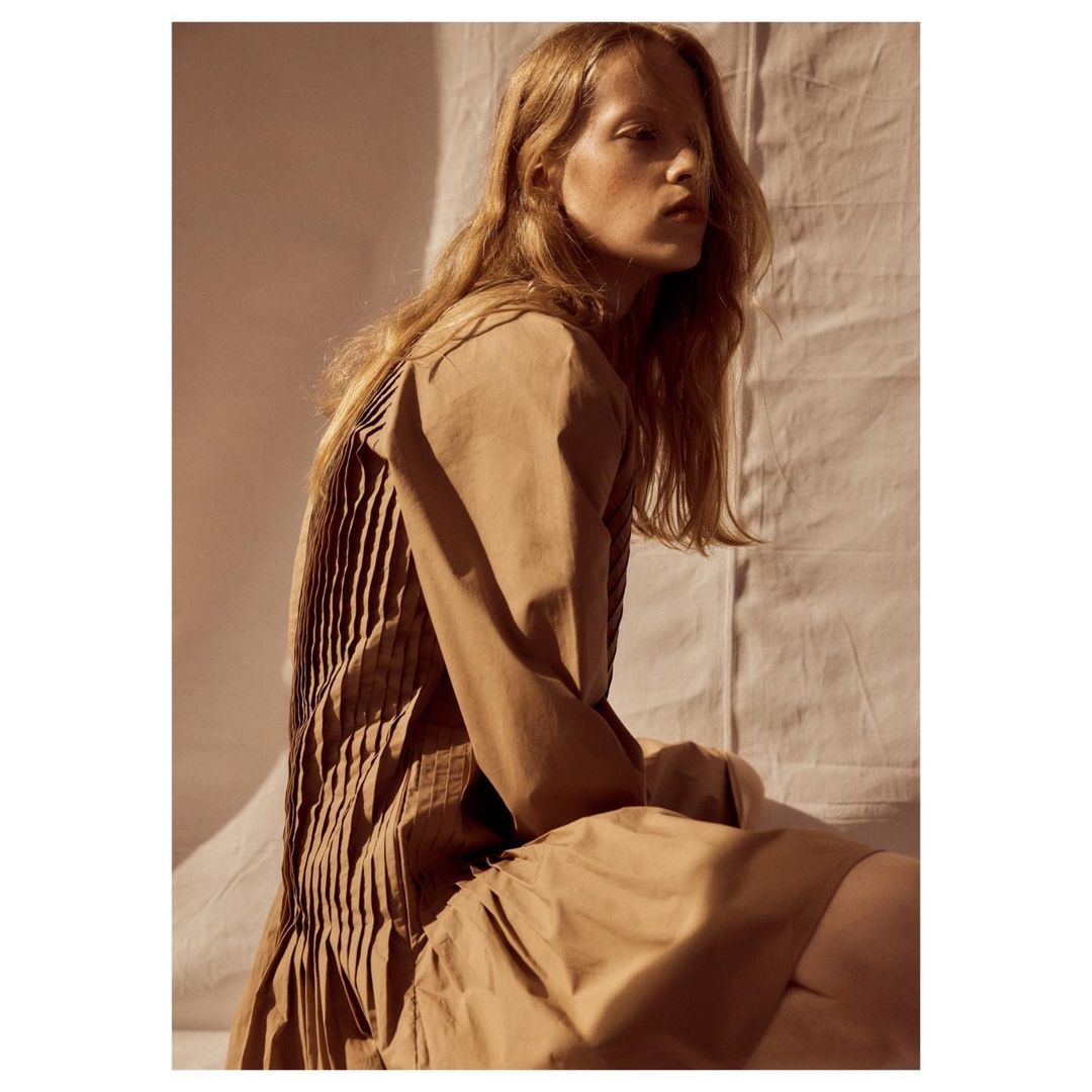 Model in a tan pleated dress seated against a wrinkled fabric background with warm sunlight